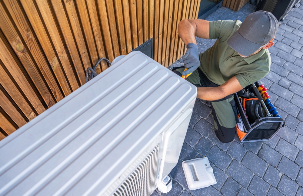 A technician works on an air conditioning unit, using tools to ensure it operates efficiently outside a contemporary building. 